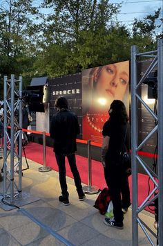 two people standing in front of a movie poster on a red carpeted area next to trees