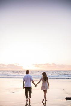 a man and woman holding hands while walking on the beach at sunset with waves coming in