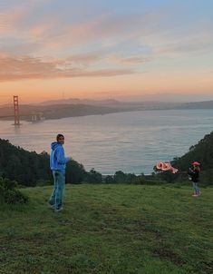 two people standing on top of a lush green hillside next to the ocean at sunset