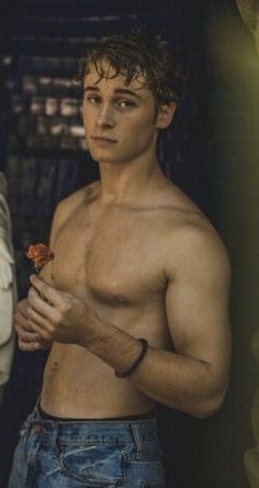 a shirtless man holding a flower in his left hand and looking at the camera