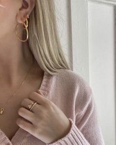 a woman wearing gold earrings and a pink sweater is holding her hand up to her chest