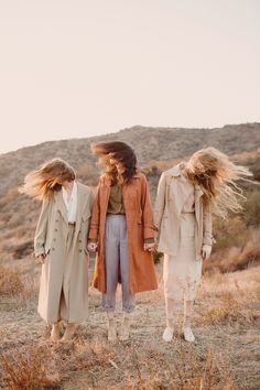 three women walking in the desert with their hair blowing in the wind and wearing trench coats