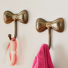 two gold bow shaped hooks hang on the wall next to a pink towel and pair of sunglasses