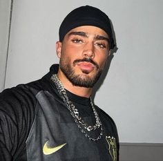 a man with a beard wearing a black shirt and gold chain necklace is standing in front of a white wall