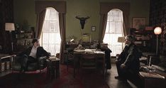 two people sitting in chairs in a living room next to windows with curtains on them