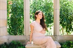 a pregnant woman sitting on a bench in front of some trees and bushes, wearing a pink dress