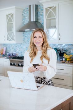 a woman standing in a kitchen holding a cell phone and looking at her laptop computer