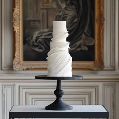 a white cake sitting on top of a black stand in front of a wall with paintings