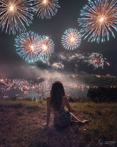 a woman sitting on top of a hill watching fireworks go off in the night sky