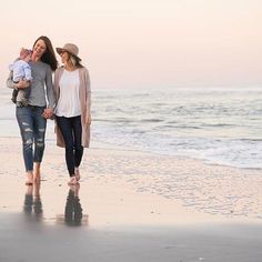 two women and a baby walking on the beach with their feet in the sand at sunset