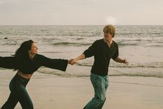 two people holding hands on the beach with waves in the background and one person running