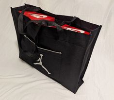 This Jordan JUMPMAN Shoe Tote Bag is perfect for those who want to keep their shoes and accessories organized while on the go. With a size of 15" x 10", this tote bag can hold up to two shoe boxes comfortably. The bag is made of high-quality canvas material with a solid black color and features a white Jumpman character on the front. It's suitable for casual occasions and has a polyester fabric type. The handle/strap is also made of canvas material. The Jordan Tote is a unisex adults' department product, with a sports theme and was manufactured in China. This tote is an excellent choice for those who want to keep their shoe collection organized and looking stylish.   This is a very sturdy and high-quality tote bag. Shoe Tote, Black Hold, Shoe Boxes, Accessory Organization, Sports Theme, Shoes And Accessories, Shoe Box, Nike Air Jordan, Canvas Material