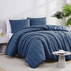 a bed with blue comforter and pillows in a room next to a plant on the floor