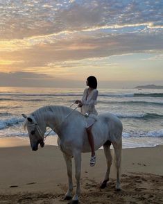 a woman riding on the back of a white horse down a beach next to the ocean