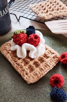 crocheted strawberries and berries are on top of a square dishcloth, next to a coffee mug