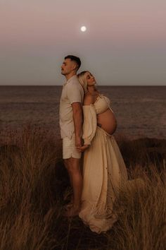 a pregnant couple standing in tall grass near the ocean