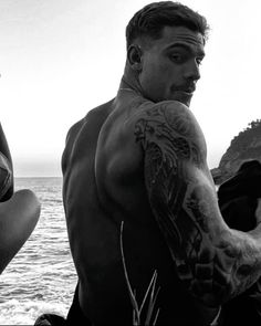 a man standing next to the ocean with tattoos on his arm