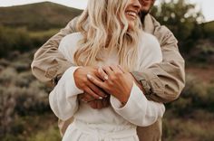 a man and woman standing next to each other in front of some bushes with their arms around each other