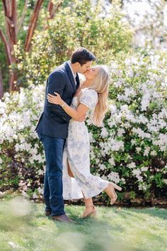 a man and woman are kissing in front of some bushes with white flowers on it