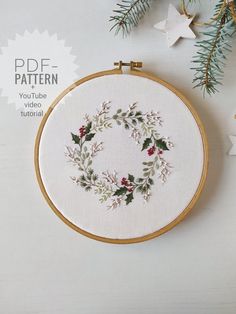 a cross stitch christmas wreath with holly and berries on white fabric surrounded by evergreen branches