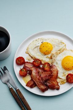 two eggs, bacon and tomatoes on a white plate next to a cup of coffee