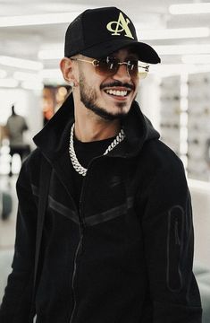 a man in a black jacket and hat smiles at the camera as he walks through an airport