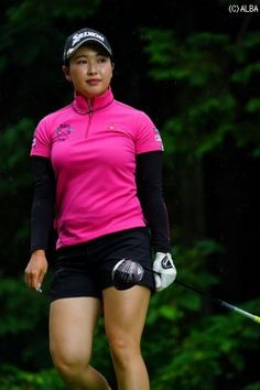 a woman in pink shirt and black shorts holding a golf club with trees in the background