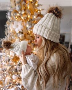 a woman holding a baby in front of a christmas tree wearing a white sweater and hat