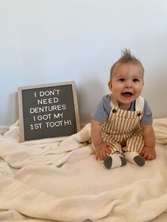 a smiling baby sitting on top of a bed next to a sign