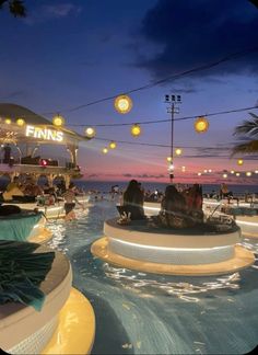 people are sitting at tables in the middle of an outdoor swimming pool with lights hanging above it