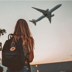 a woman with a backpack looking at an airplane in the sky over her head,