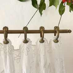 the curtain rod is holding two white curtains with gold hardware and flowers on them, along with green leaves