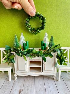 a hand reaching for a wreath on top of a white table with green trees and candles