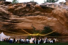 a group of people sitting at tables in front of a rock formation with lights strung from it