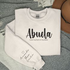 This sweatshirt is a heartwarming tribute to the cherished bond between Spanish grandmothers and their beloved grandkids. This sweater beautifully weaves together the essence of an Abuela's love and the joy of proudly showcasing the names of her precious grandchildren. SHIRT INFORMATION AND MATERIAL Crafted from 50% Cotton and 50% Polyester, our sweatshirt offers superior comfort and durability. Its relaxed fit and durability make it perfect for long days, family outings, or simply running erran Heart On My Sleeve, Grandma Sweatshirt, Family Outings, Custom Hoodie, Personalized Grandma, Family Outing, Small Business Ideas, Custom Hoodies, Heart On