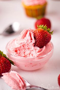 two strawberries sit in a bowl with pink frosting on the table next to cupcakes