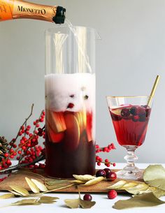 two glasses filled with liquid sitting on top of a table next to red berries and gold leaves