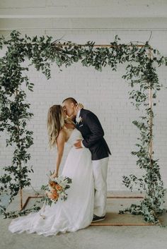 a bride and groom kissing in front of an arbor with greenery on the wall