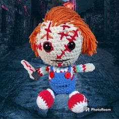 a crocheted doll with red hair and big eyes sitting in an alleyway