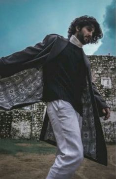 a man walking across a dirt field next to a stone wall with a long coat over his shoulders