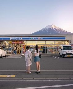 two women are standing in front of a store with cars parked outside and a mountain in the background