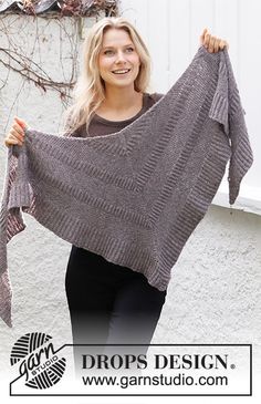 a woman holding up a gray shawl in front of a white building with the words drops design on it