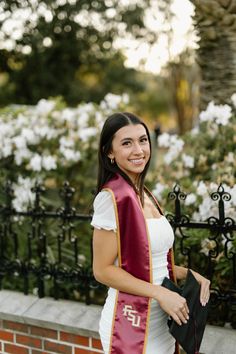 a woman in a red and white graduation gown standing next to a brick wall with flowers behind her