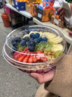 a person is holding a plastic bowl full of fresh fruit and cereal on the street