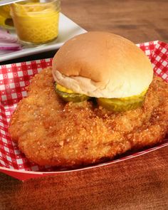 a fried chicken sandwich with pickles and mustard on a red checkered paper tray