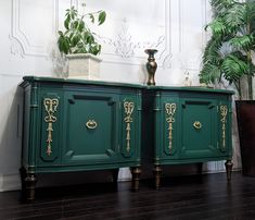 two green sideboards with gold handles on them in front of a white wall and potted plant