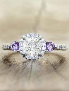 a diamond and amethyst engagement ring on a wooden surface with other jewelry items in the background