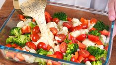 a person pouring dressing onto a salad in a glass dish with broccoli, tomatoes and cauliflower