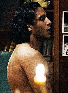 a man with no shirt on standing in front of a book shelf holding a lit candle