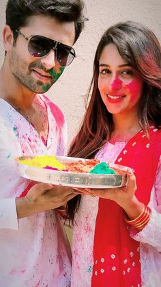 a man and woman standing next to each other holding a plate with colored powder on it